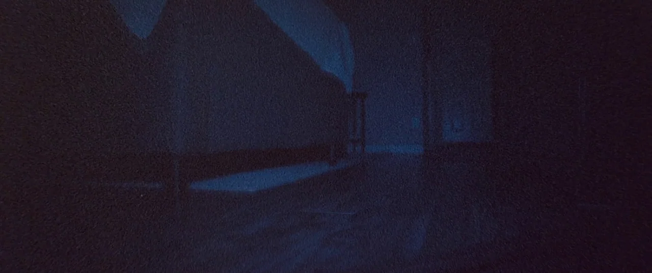 Skinamarink (2022) movie still: establishing — A dark bedroom interior is illuminated by a faint blue light source. A bed frame rests against the…