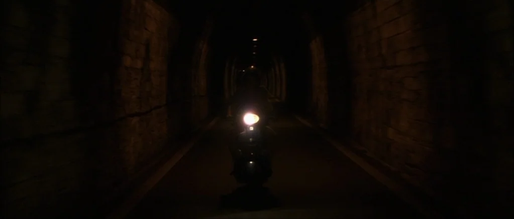 Genova (2008) movie still: wide — A dark, arched stone tunnel extends into the distance with rough wall textures. A lone motor scooter drives…