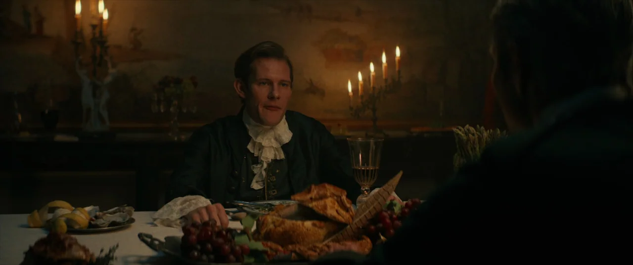 The Promised Land (2023) movie still: medium — A dimly lit 19th-century dining table with a spread of food and grapes. A man sits at the head of the…
