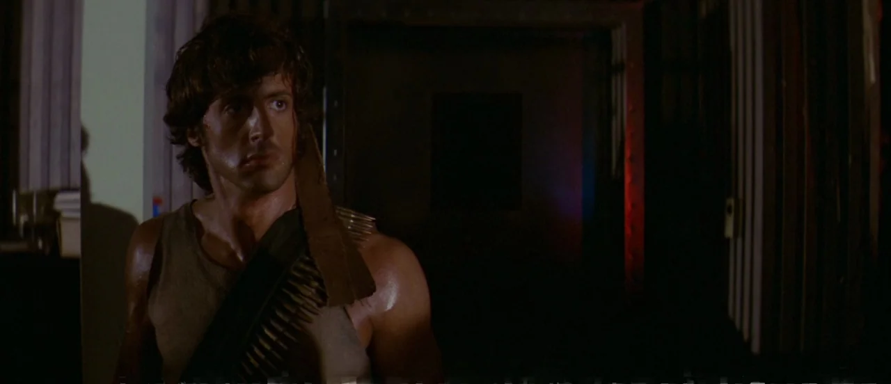 First Blood (1982) movie still: medium — A man stands in a shadowed, dimly lit corridor with metallic door frames. He looks off to the side while…