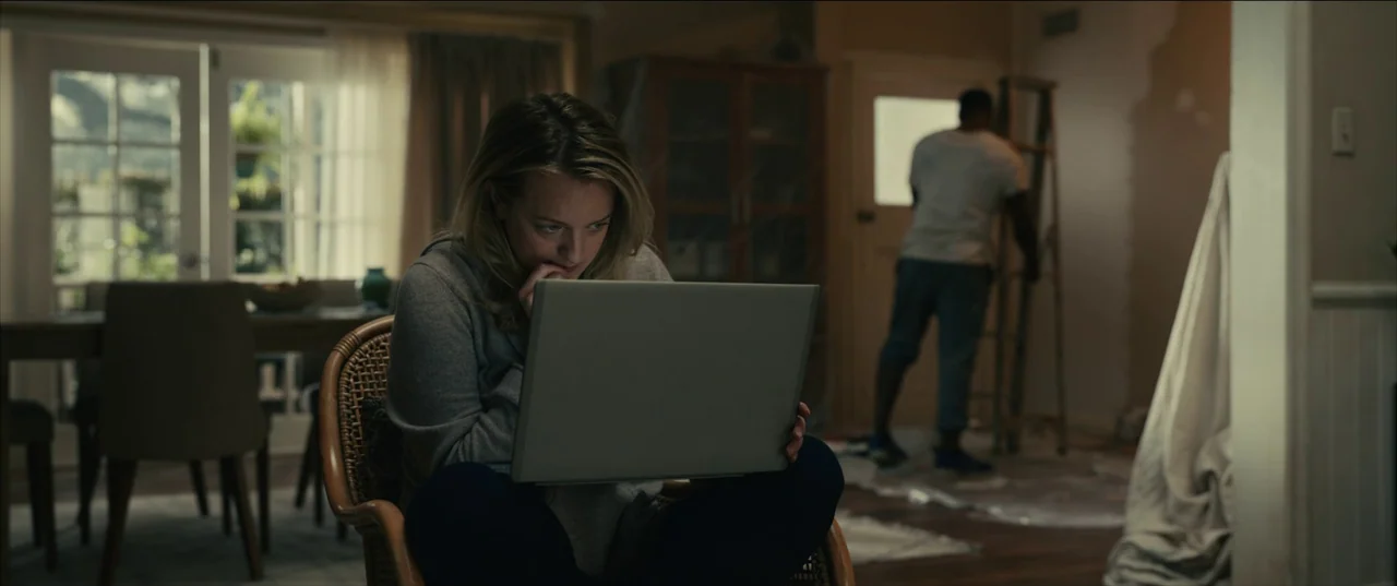The Invisible Man (2020) (2020) movie still: medium — A woman sits in a wicker chair working on a laptop within a home. Behind her a man climbs a…