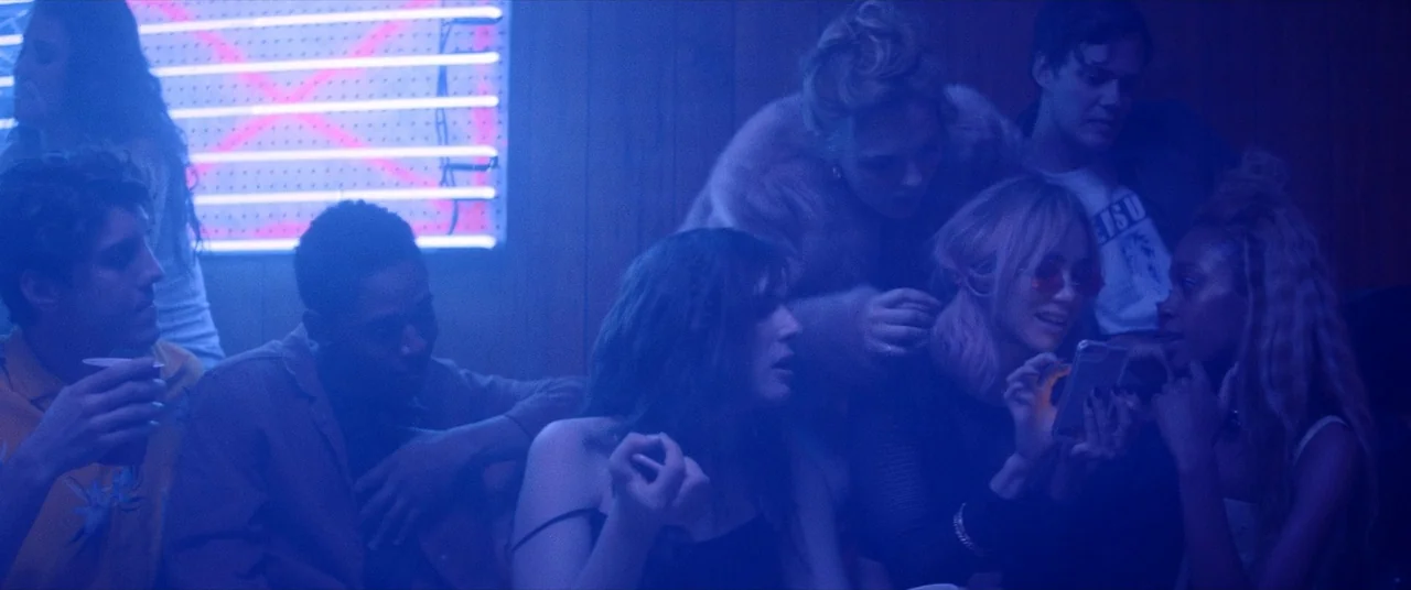 Assassination Nation (2018) movie still: medium — A dimly lit room with wood-paneled walls and a bright horizontal neon sign fixture. Several young…