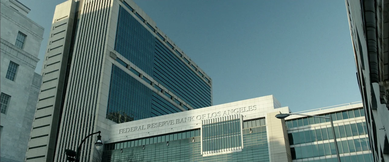 Den of Thieves (2018) movie still: wide — A modern commercial building facade with glass windows and prominent stone lettering at street level. The…