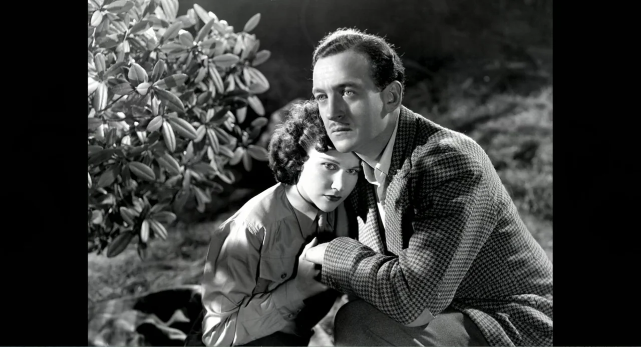 Made in England: The Films of Powell and Pressburger (2024) movie still: medium — A man and woman crouch together outdoors near a large leafy shrub.…