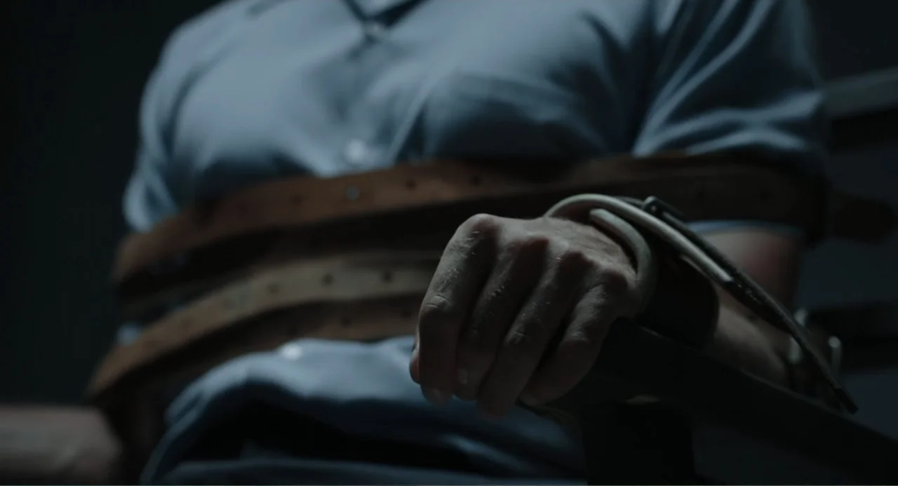 Ted Bundy: American Boogeyman (2021) movie still: closeup — A dimly lit room focuses on a person strapped to a chair with brown leather belts. The…