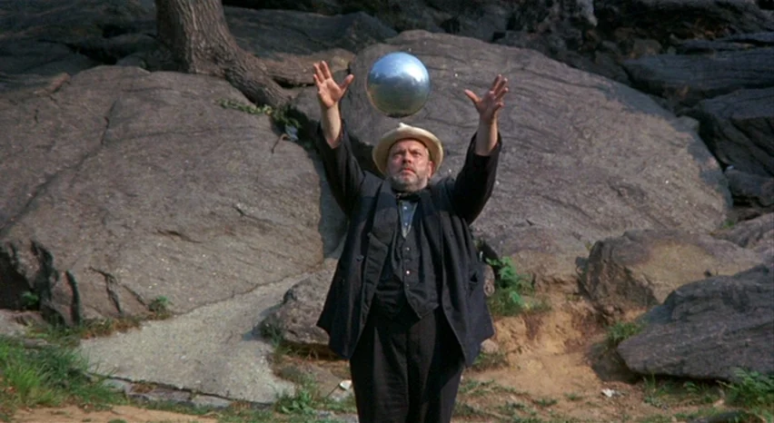 A Safe Place (1971) movie still: medium — A man stands before a massive, weathered grey rock formation in a wooded area. He reaches both arms upward…