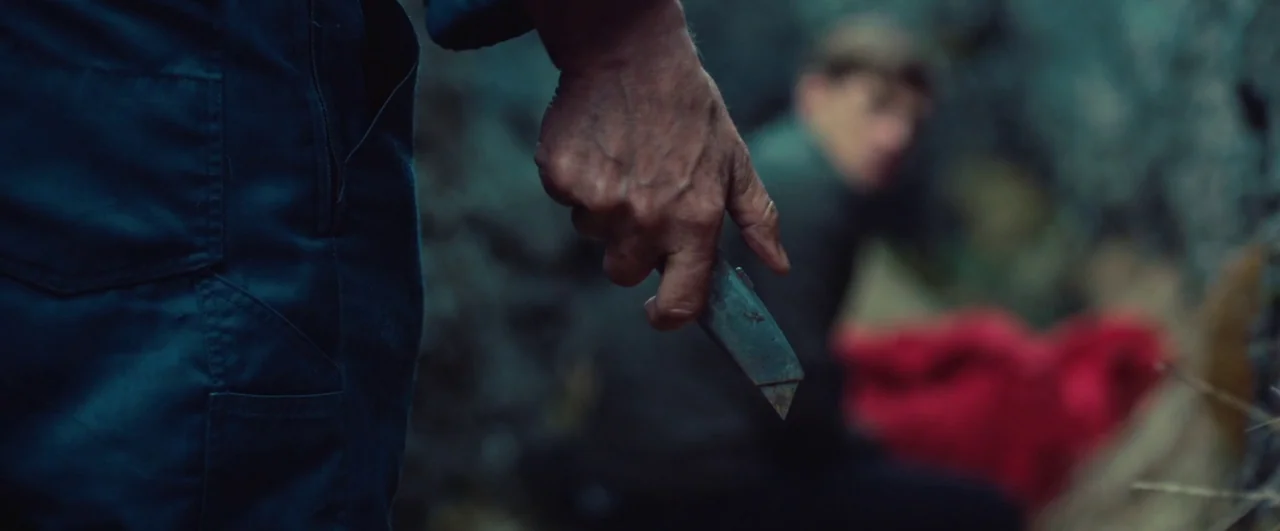 Rift (2017) movie still: closeup — A close-up shot focuses on a weathered hand gripping a small utility knife blade. A person is crouched in the…