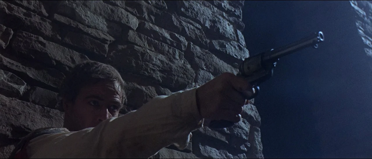 The Appaloosa (1966) movie still: medium — A man stands before a rough-hewn stone wall in near darkness. He holds a metal revolver extended toward…