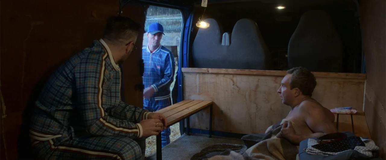 The Gentlemen (2020) movie still: medium — The interior of a cargo van features plywood paneling and a wooden bench. Two men in matching plaid…