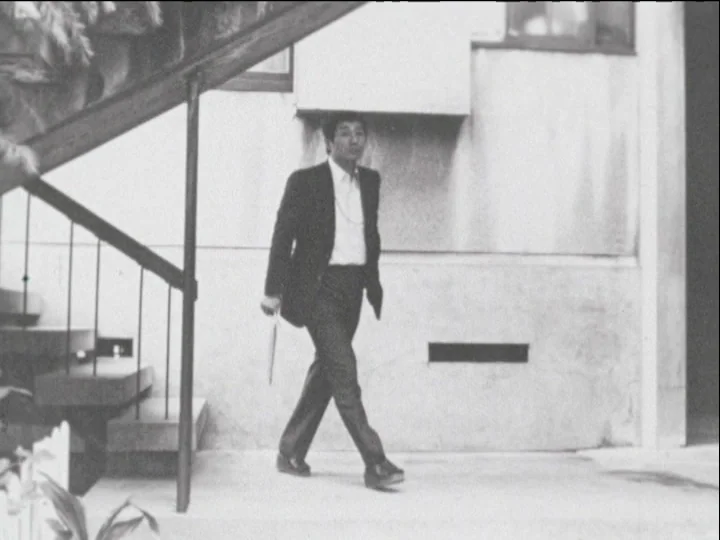 Catalogue of Memory (1977) movie still: fullbody — An exterior architectural space features a concrete wall and a metal staircase structure. A man…
