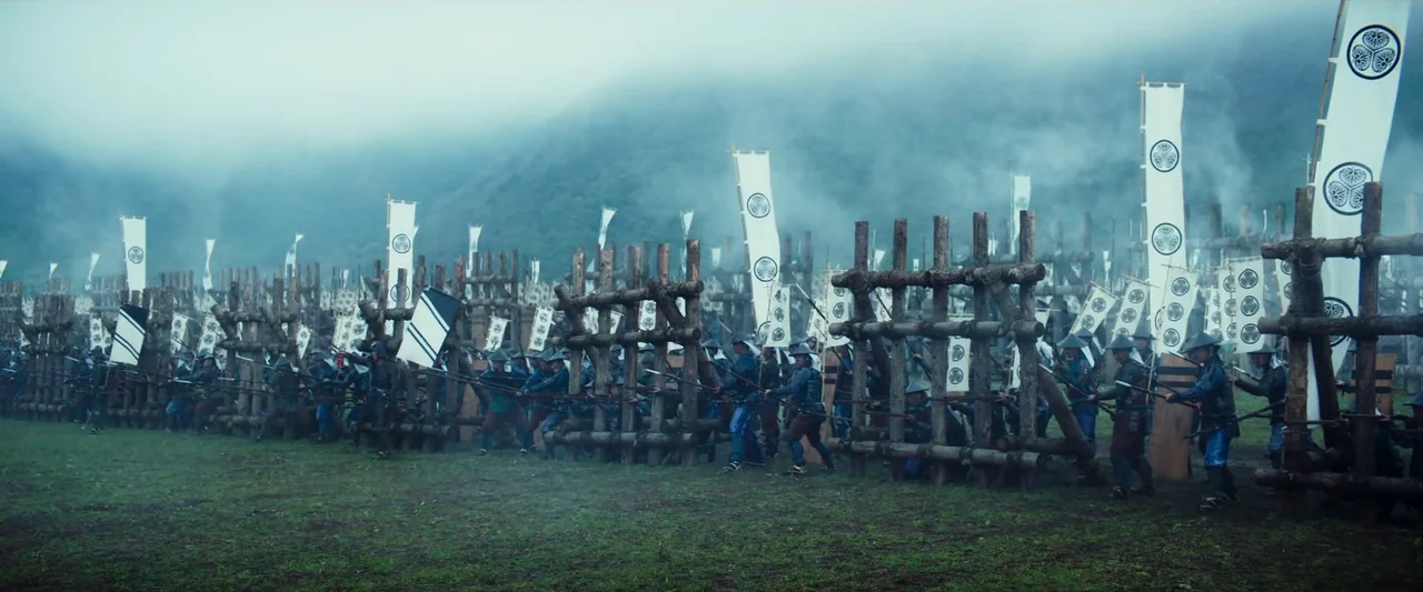 Kubi (2023) movie still: wide — A line of soldiers behind wooden defensive barricades waits on a grassy battlefield. Fog rolls over the distant…