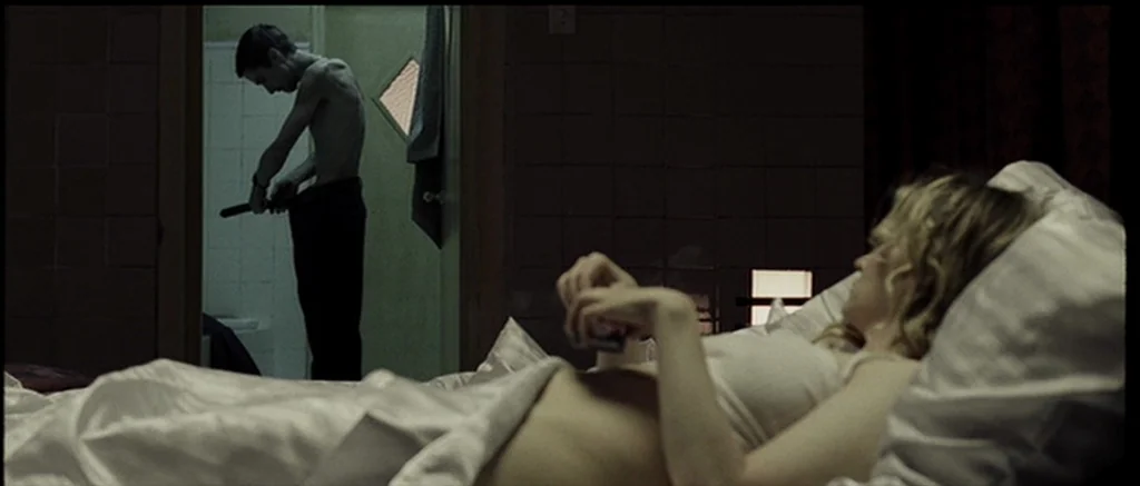 The Machinist (2004) movie still: wide — A dimly lit room with an adjoining bathroom visible through an open door. An extremely thin man buckles a…