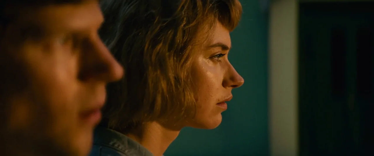 Vivarium (2019) movie still: closeup — A dark indoor setting is lit by a focused warm light source. A woman in profile stares ahead while a man is…
