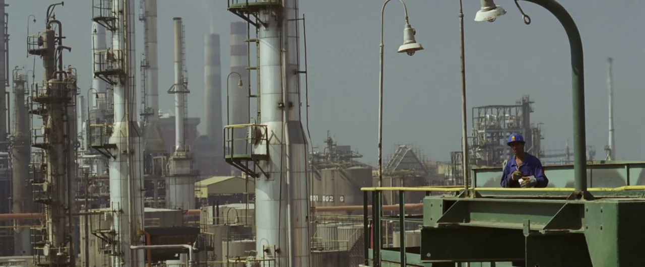 Syriana (2005) movie still: wide — An sprawling oil refinery features tall silver distillation towers and complex piping networks. A lone worker…