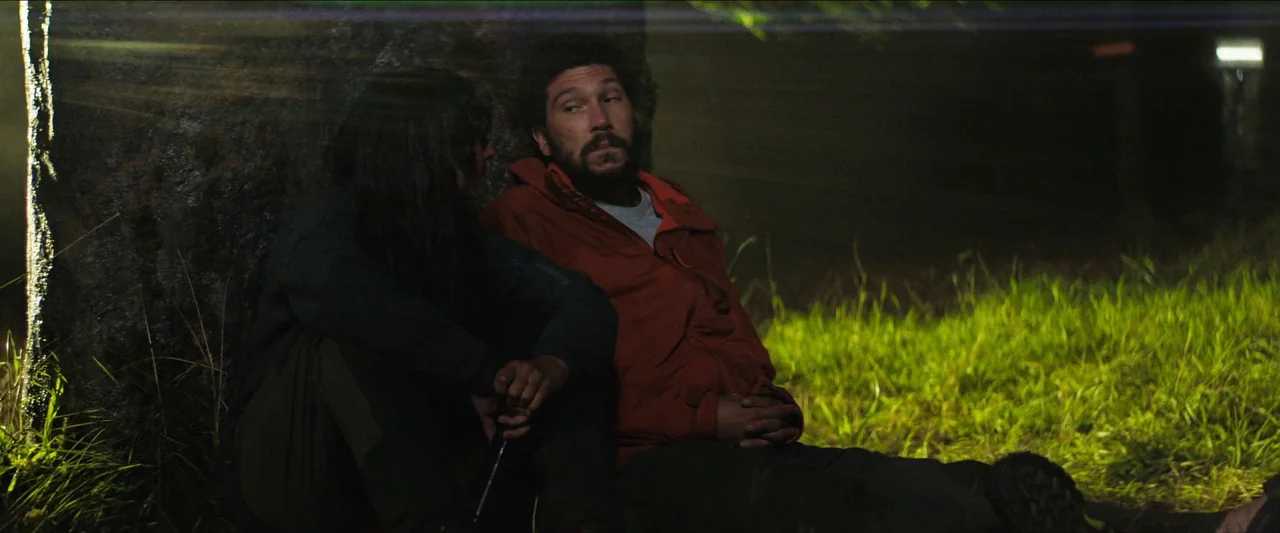 In The Earth (2021) movie still: medium — Two individuals sit on a dark grassy incline backed by a large tree trunk. The pair rests against the wood…