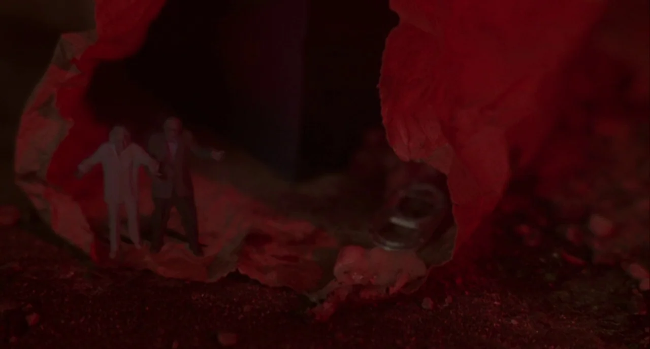 Mulholland Drive (2001) movie still: closeup — Two miniature human figurines stand on crumpled red paper next to a soda tab. The scene is lit with…