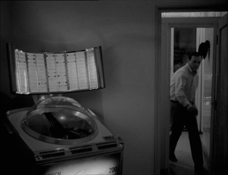 Alphaville (1965) movie still: medium — A dark, dimly lit room contains a large vintage jukebox in the foreground. A man walks through a doorway…