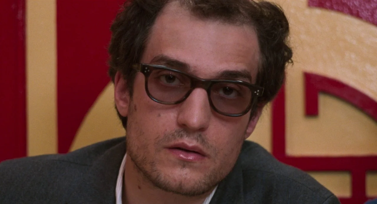Godard Mon Amour (2015) movie still: closeup — A man sits in front of a red and gold decorative wall pattern. The subject stares straight ahead at…