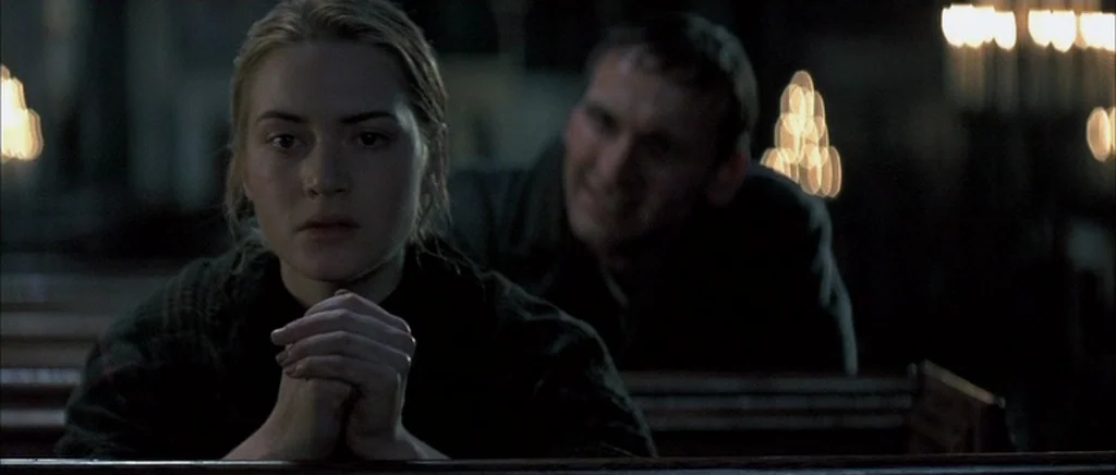 Jude (1996) movie still: medium — A dimly lit, wooden-paneled space with rows of pews and glowing candle flames. A young woman with her hands…