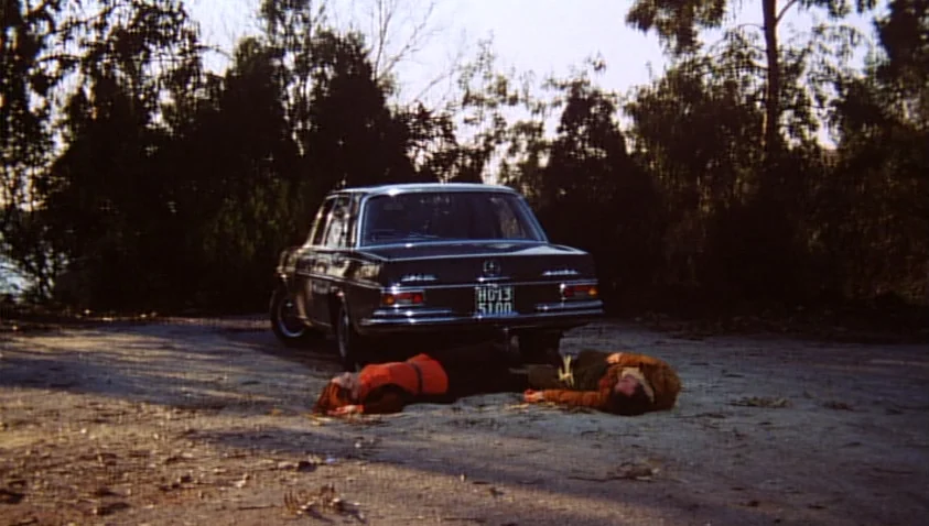 A Bay of Blood (1971) movie still: wide — A dark vintage sedan is parked in a wooded area with dense foliage behind it. Two figures are lying prone…