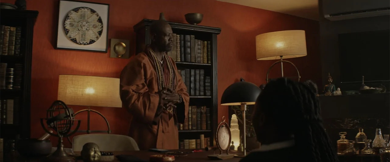 A Scam Called Love (2025) movie still: medium — A study features dark wooden bookshelves filled with books against a deep orange wall. A bald man in…