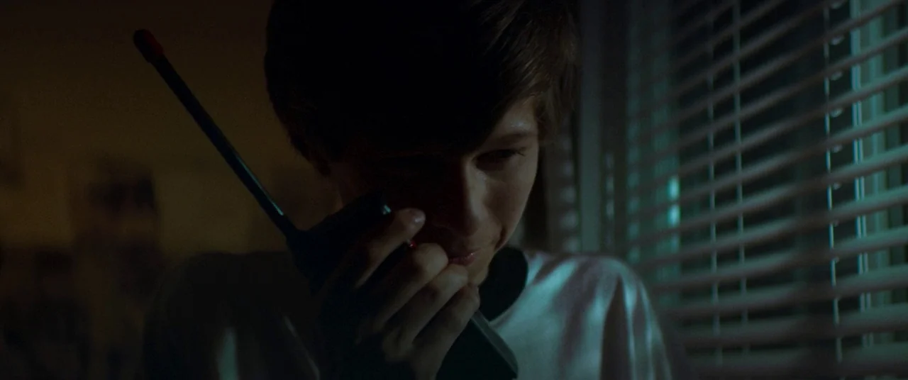 Summer of 84 (2018) movie still: closeup — A dimly lit room features a window with partially closed horizontal blinds. A teenage boy stands near the…