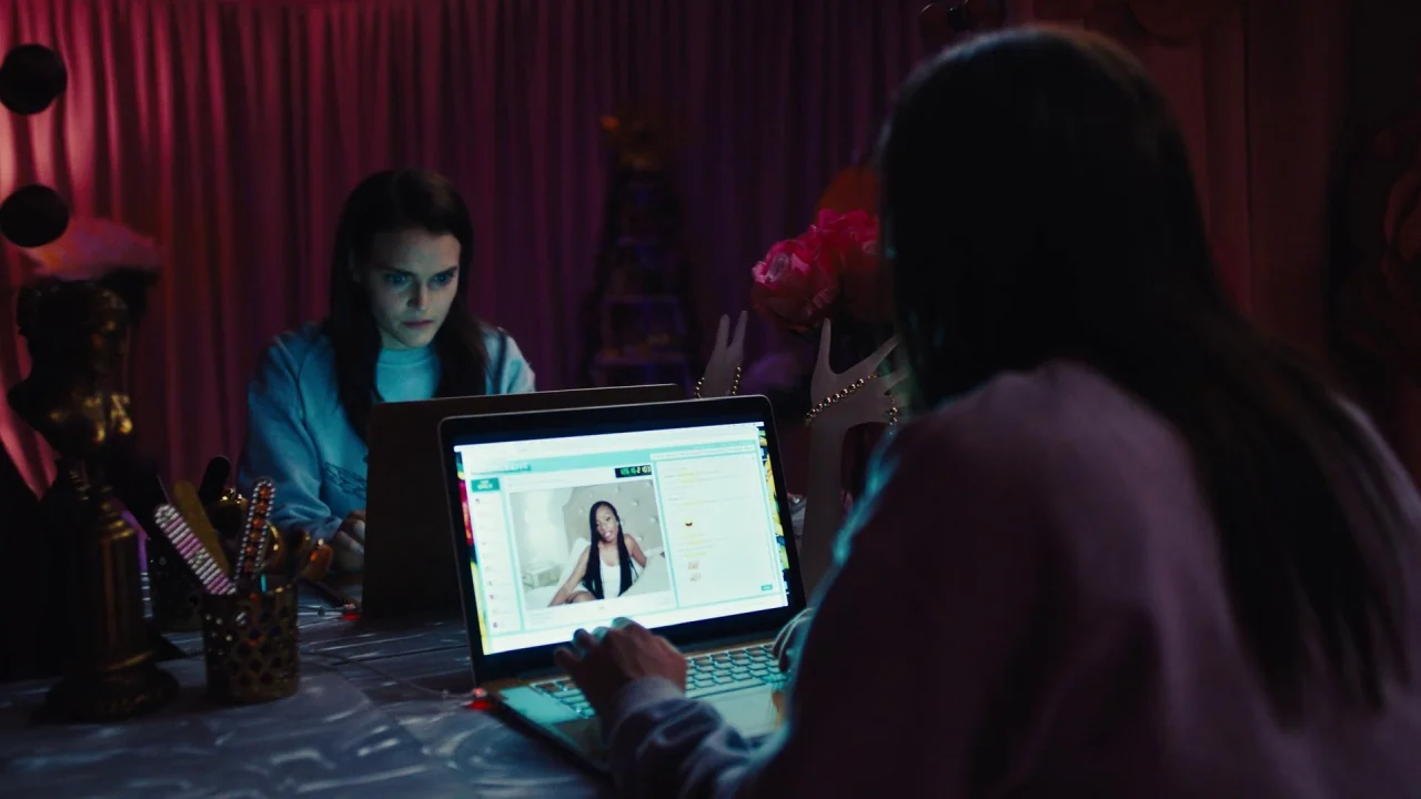 Cam (2018) movie still: medium — A dimly lit room with draped pink fabric serves as a makeshift workstation. Two women sit at a table illuminated by…