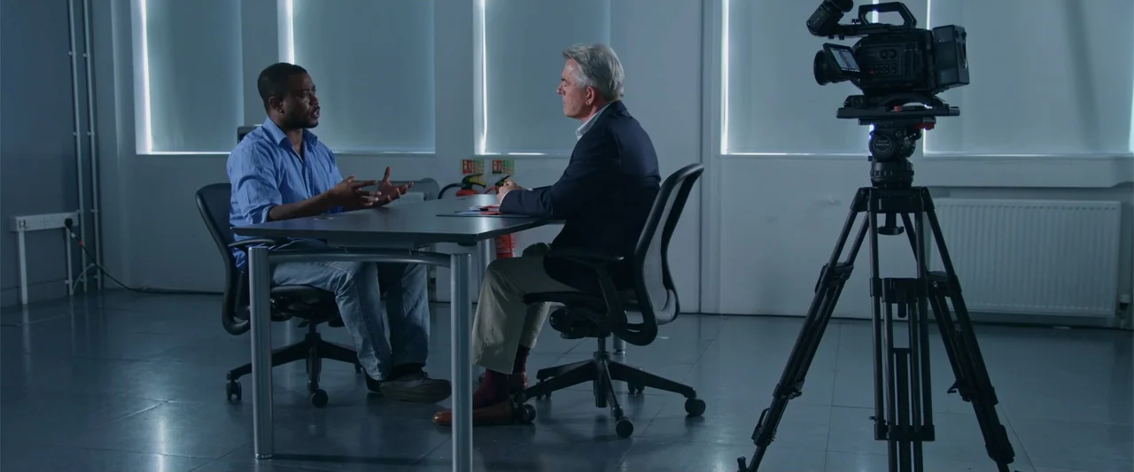 Beyond the Universe (2022) movie still: medium — A spartan room features a desk and office chairs positioned before large windows. Two men sit…