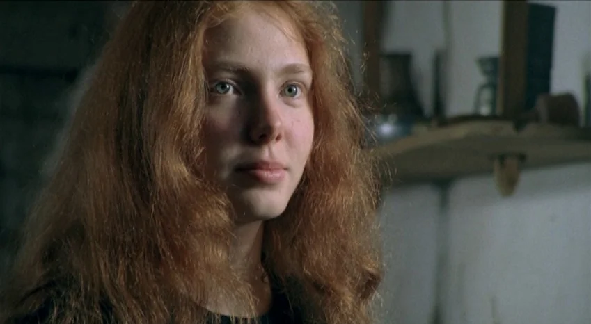 Bluebeard (2009) movie still: closeup — A dimly lit indoor space with white walls and a wooden shelf behind. A young woman with long auburn hair…