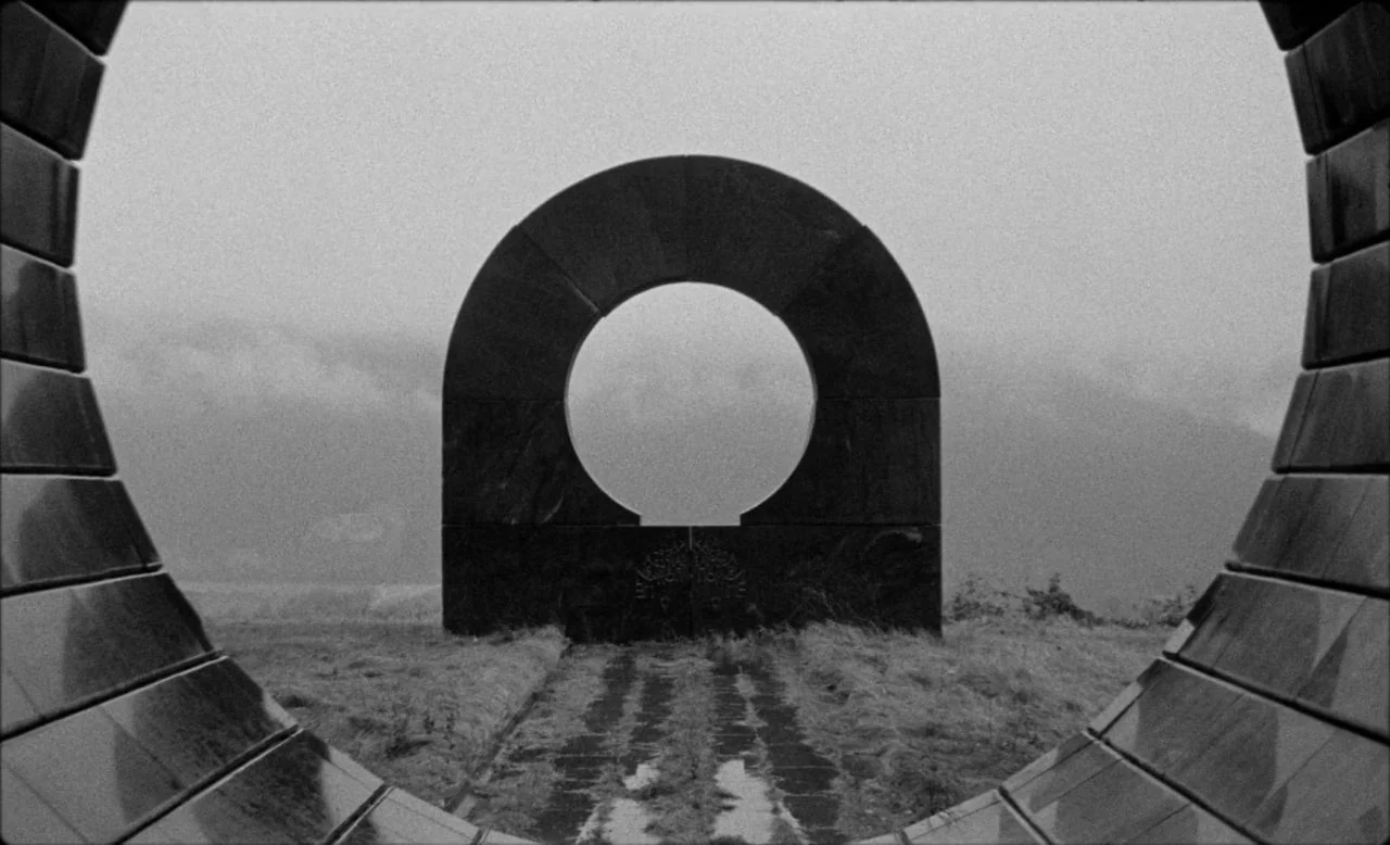 Last and First Men (2020) movie still: establishing — A stone monument with a central circular opening stands in an open grassy field. A stone-paved…