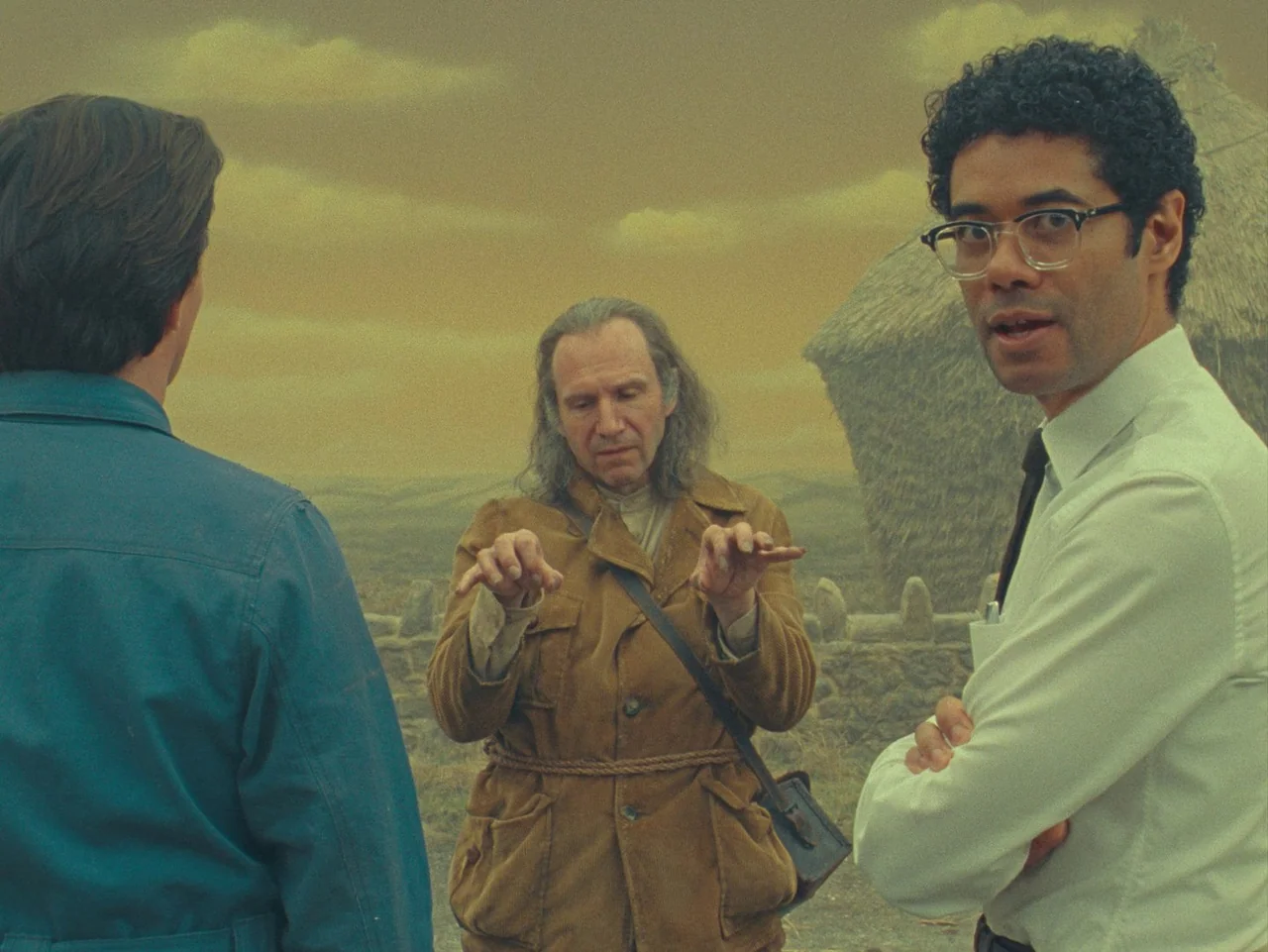 The Rat Catcher (2023) movie still: medium — A man stands between two others in a flat landscape under a hazy sky. The central man makes hand…
