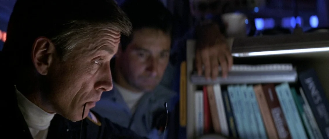 The Hunt for Red October (1990) movie still: closeup — A dimly lit interior space with blue lighting and a shelf of reference books. Two men in…