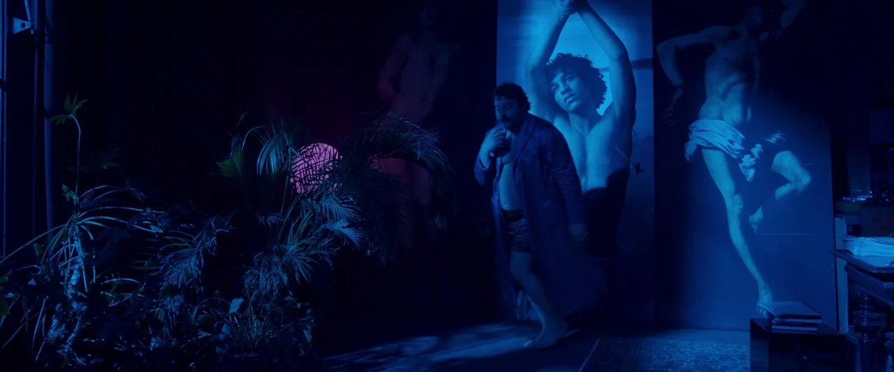Peter Von Kant (2022) movie still: fullbody — A dark space features large vertical wall prints of male figures in blue light. A heavy-set man in a…