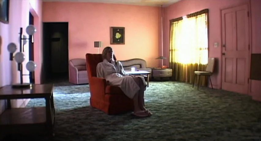Inland Empire (2006) movie still: wide — A spacious room features pink walls, green carpet, and mid-century furniture pieces. A woman in a white…