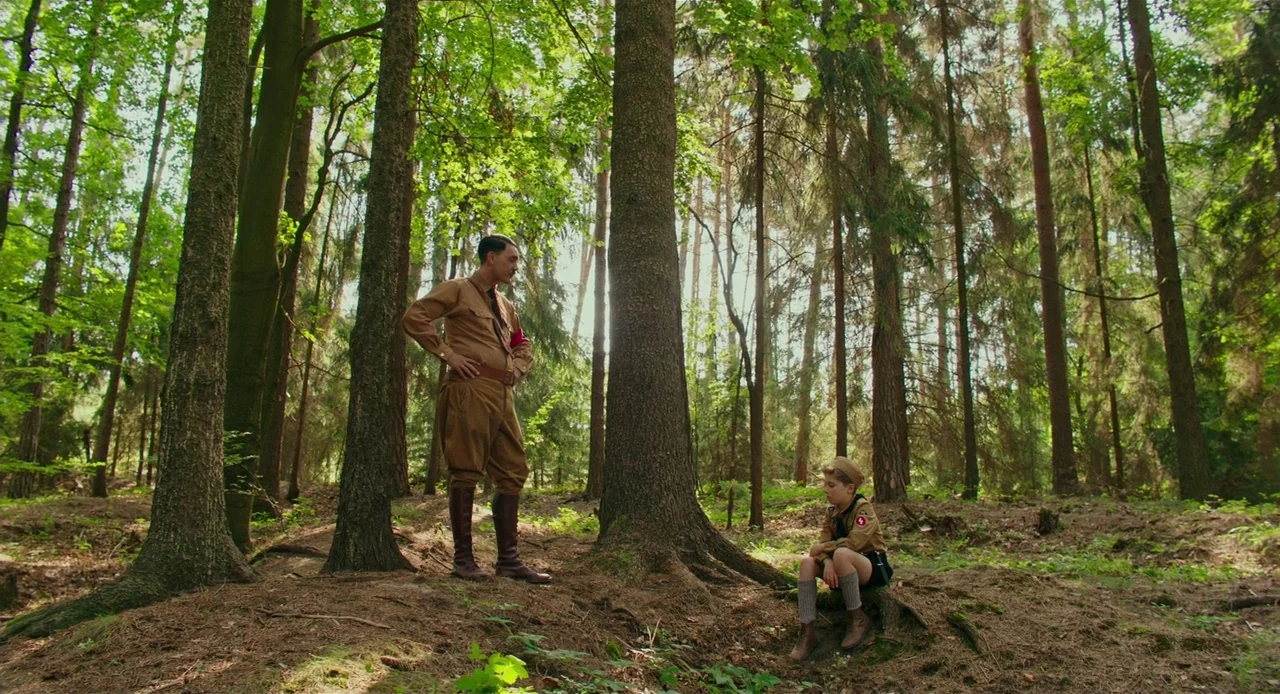 Jojo Rabbit (2019) movie still: fullbody — A dense woodland area with tall pine trees and a mossy, needle-covered floor. A man in historical uniform…