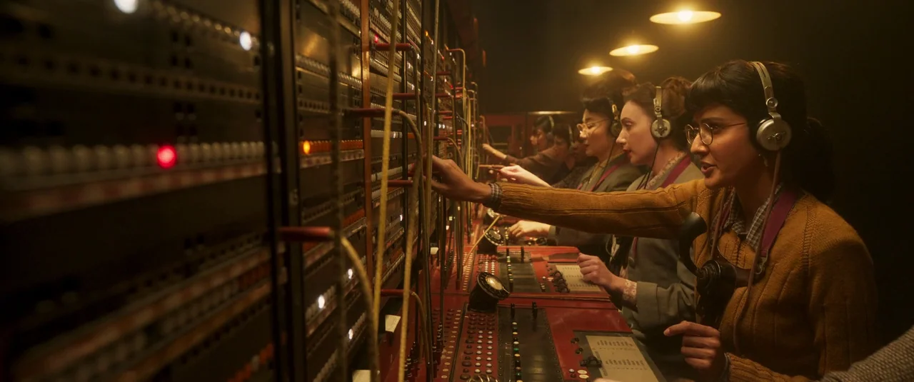 Wonka (2023) movie still: medium — A dark, wood-paneled telephone exchange room houses multiple operators at a long control desk. Several women wear…