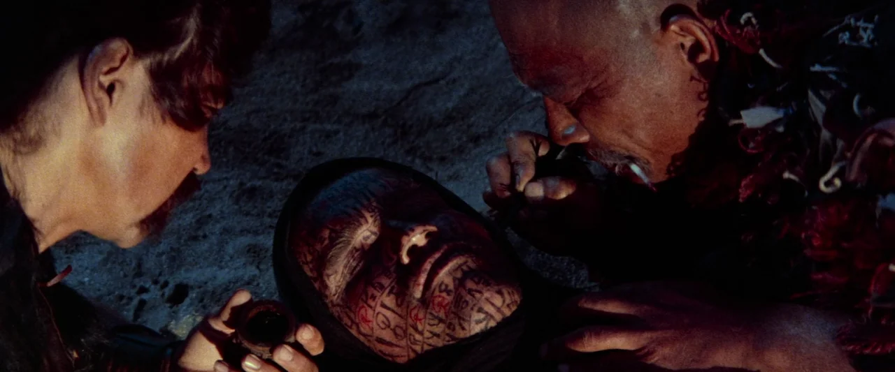 Conan The Barbarian (1982) movie still: medium — A dark, sandy exterior at night lit by a flickering warm light source. Two men lean over a third…