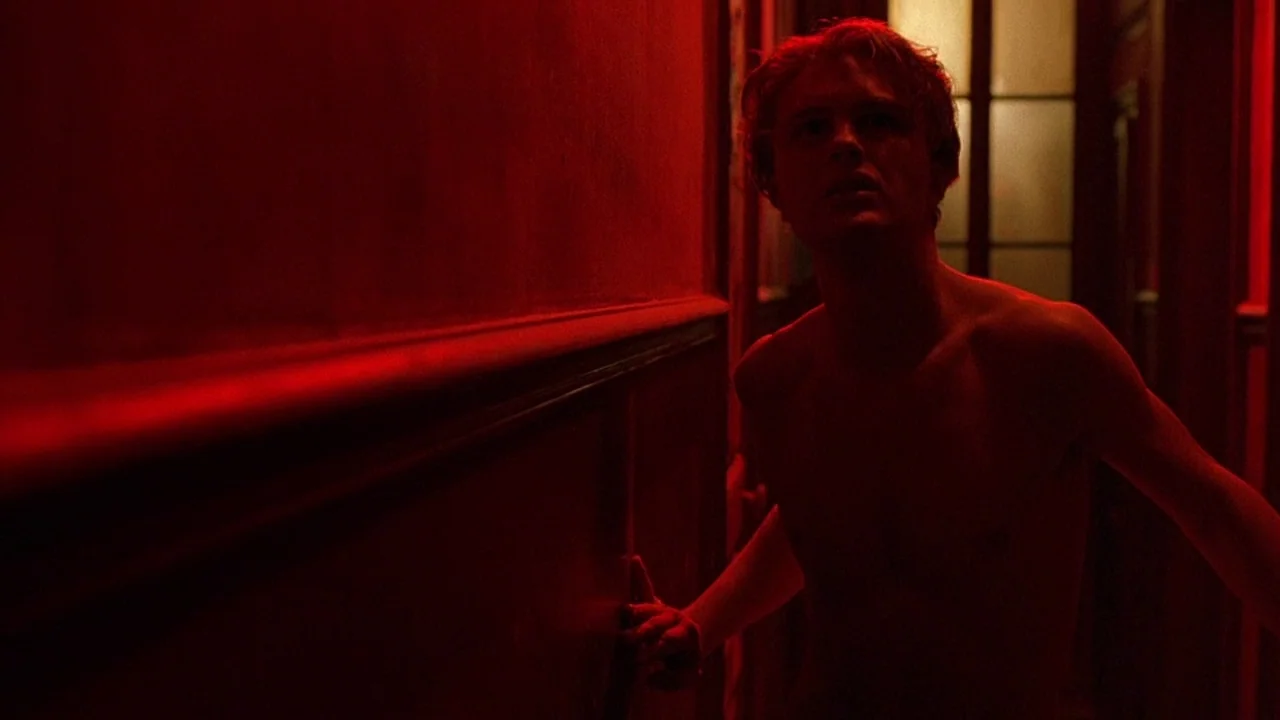 The Dreamers (2003) movie still: medium — A narrow residential hallway is drenched in deep red light from an unseen source. A shirtless man stands…