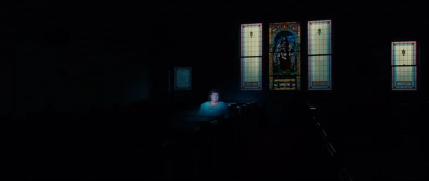 Bubble (2005) movie still: wide — A dimly lit church interior with rows of empty wooden pews in deep darkness. A lone woman sits centered in the…