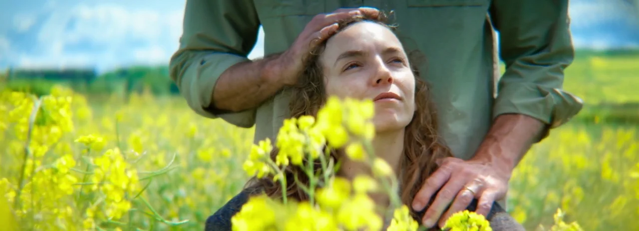 28 Years Later (2025) movie still: medium — A field of vibrant yellow flowers blooms under a bright sky. A man stands behind a woman, gently resting…