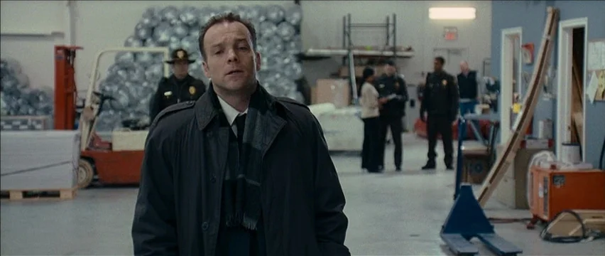 Snow Angels (2007) movie still: medium — A wide, sparse industrial facility features stacks of wrapped materials and metal equipment. Several men in…