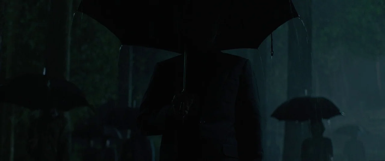Satan’s Slaves (2017) movie still: medium — A dark, rainy woodland setting at night contains several figures holding umbrellas. The central figure…