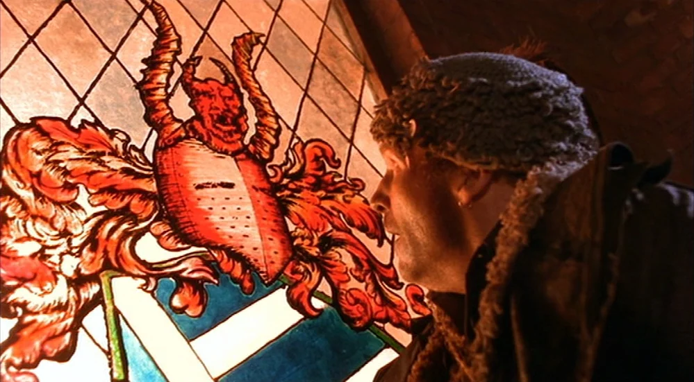 The Fisher King (1991) movie still: medium — A man stands before a large stained glass window depicting a red horned crest. The man in a fur-lined…