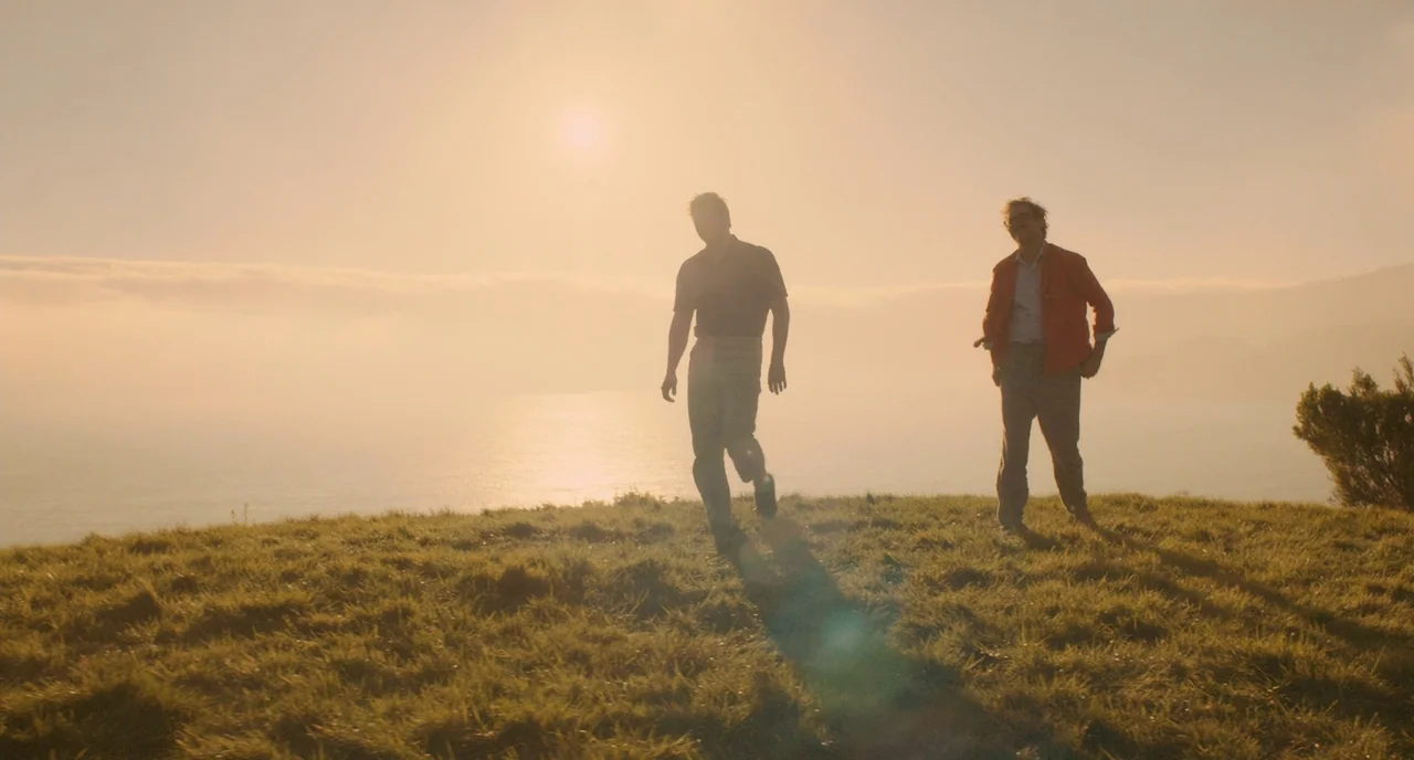 Her (2013) movie still: fullbody — A grassy hill overlooks the ocean at sunset with hazy light conditions. Two men walk across the hill while the…