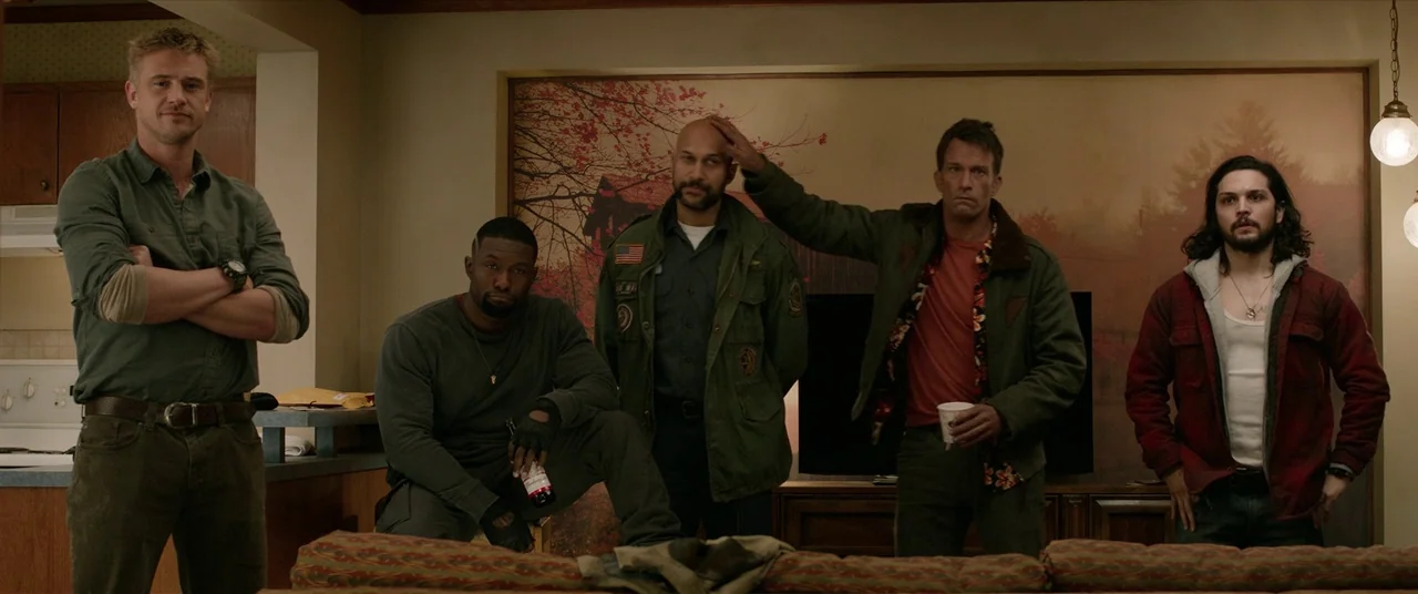 The Predator (2018) movie still: fullbody — A residential living room features an autumnal forest mural on the back wall. Five men stand together in…