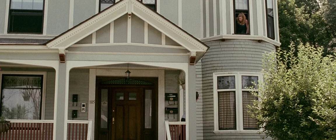 The Fighter (2010) movie still: wide — A grey-sided suburban house with white trim features a wooden front door. A young woman looks out from an…
