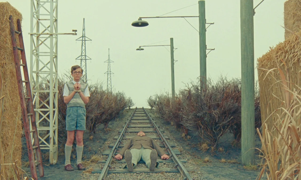 The Swan (2023) movie still: wide — A desolate railway line stretches through a flat landscape filled with dry, leafless bushes. A young boy stands…