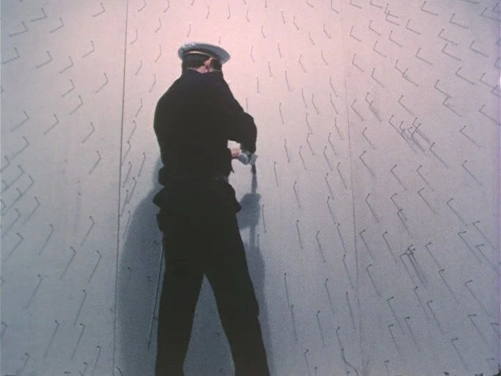 Trial (1975) movie still: fullbody — A man in a dark uniform faces a plain white wall covered in patterns. He is in the process of affixing small,…