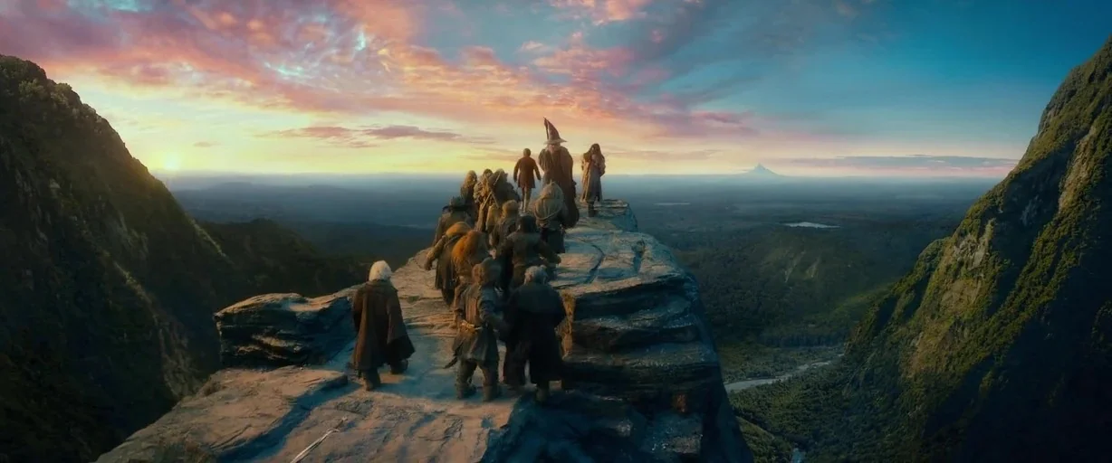 The Hobbit: An Unexpected Journey (2012) movie still: wide — A group of travelers walks along a stone cliff edge at sunset. The high mountain path…