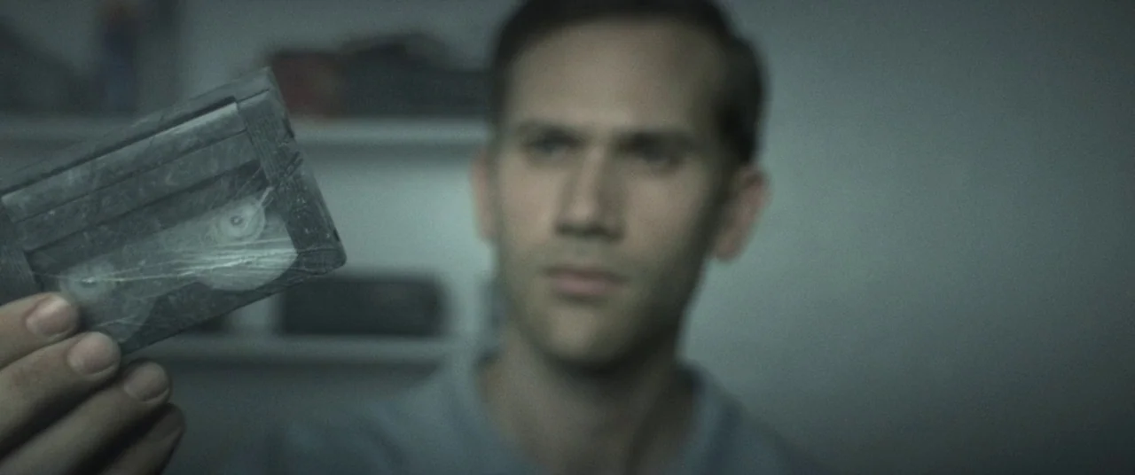 The Endless (2017) movie still: closeup — A dimly lit room with blurred shelves visible in the background. A man holds a small plastic cassette tape…